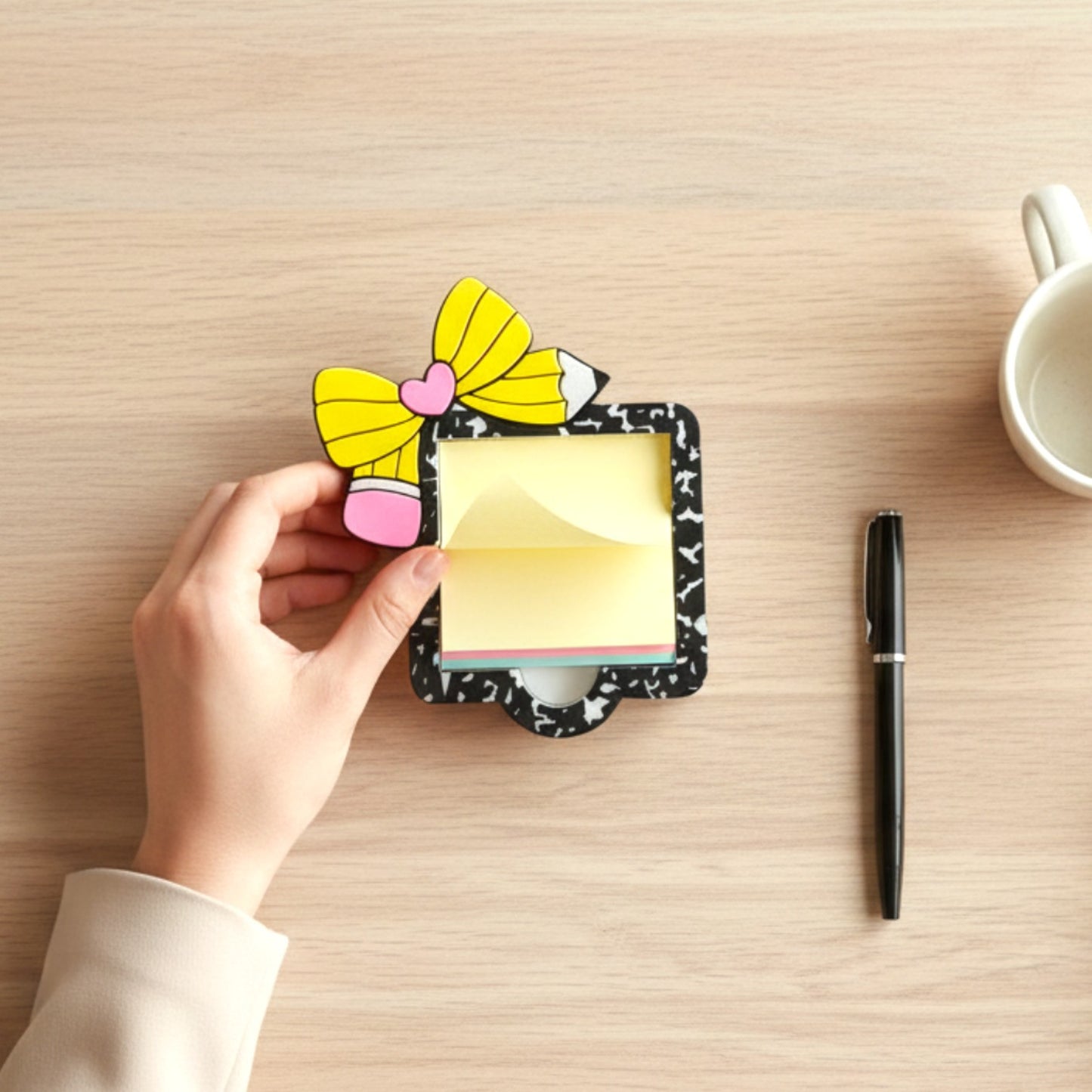 Xpress It Graphics yellow pencil and pink bow sticky note holder with black speckled frame, held by a person in a beige blazer.